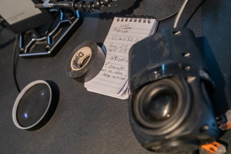 Scattered items lie on a black desk: a roll of electrical tape, a glass lens, components from a camera, and a notepad containing numbers and the heading 'LLM'. Other miscellaneous items of hardware are out of focus in the background.